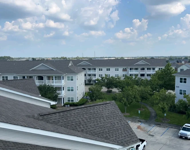 Elevated view of multi-story apartment buildings with dark shingled roofs surrounding a landscaped courtyard and parking area.